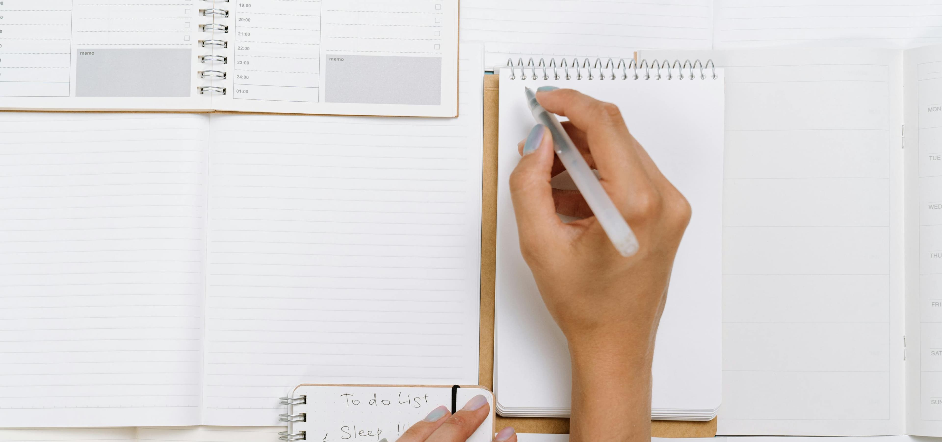 A hand writes in a spiral notebook, with open planners and a to-do list, evoking a sense of organization.