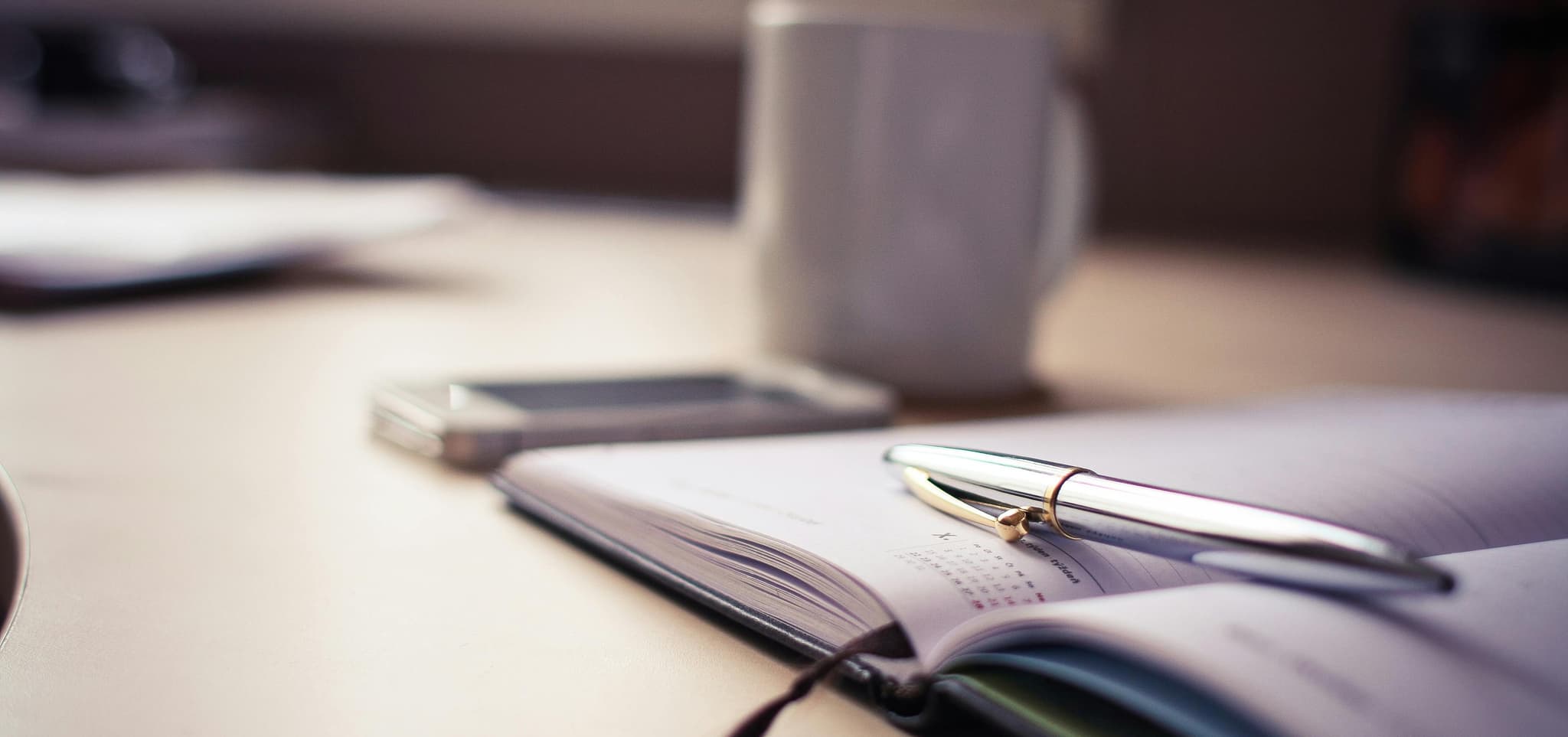 A silver pen on an open planner with dated pages, beside a blurred smartphone and mug, conveying productivity.