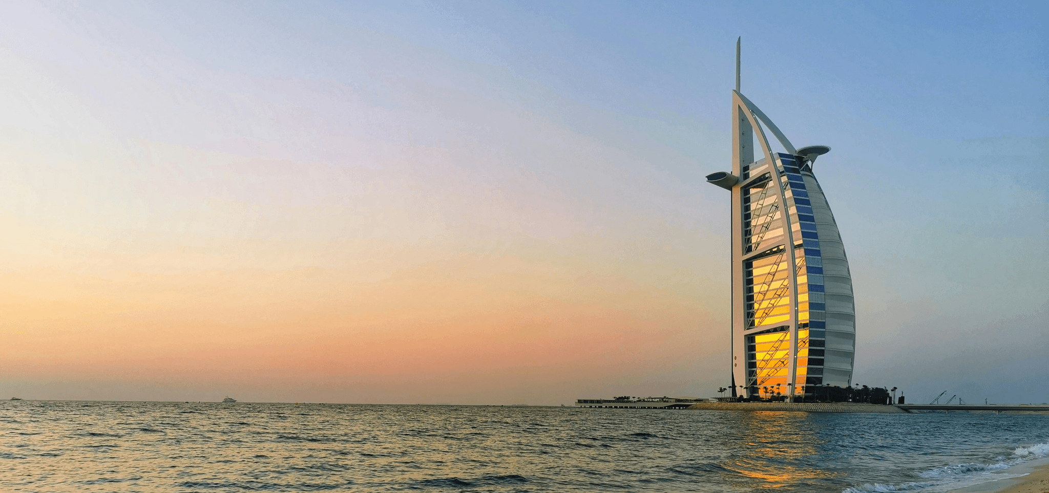 Sunset over the ocean with a sail-shaped skyscraper reflecting golden light, evoking a serene atmosphere.