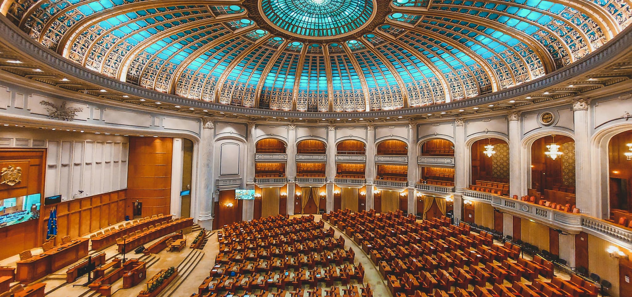 Ornate parliament hall with a blue stained-glass dome ceiling and rows of wooden seats, exuding grandeur.