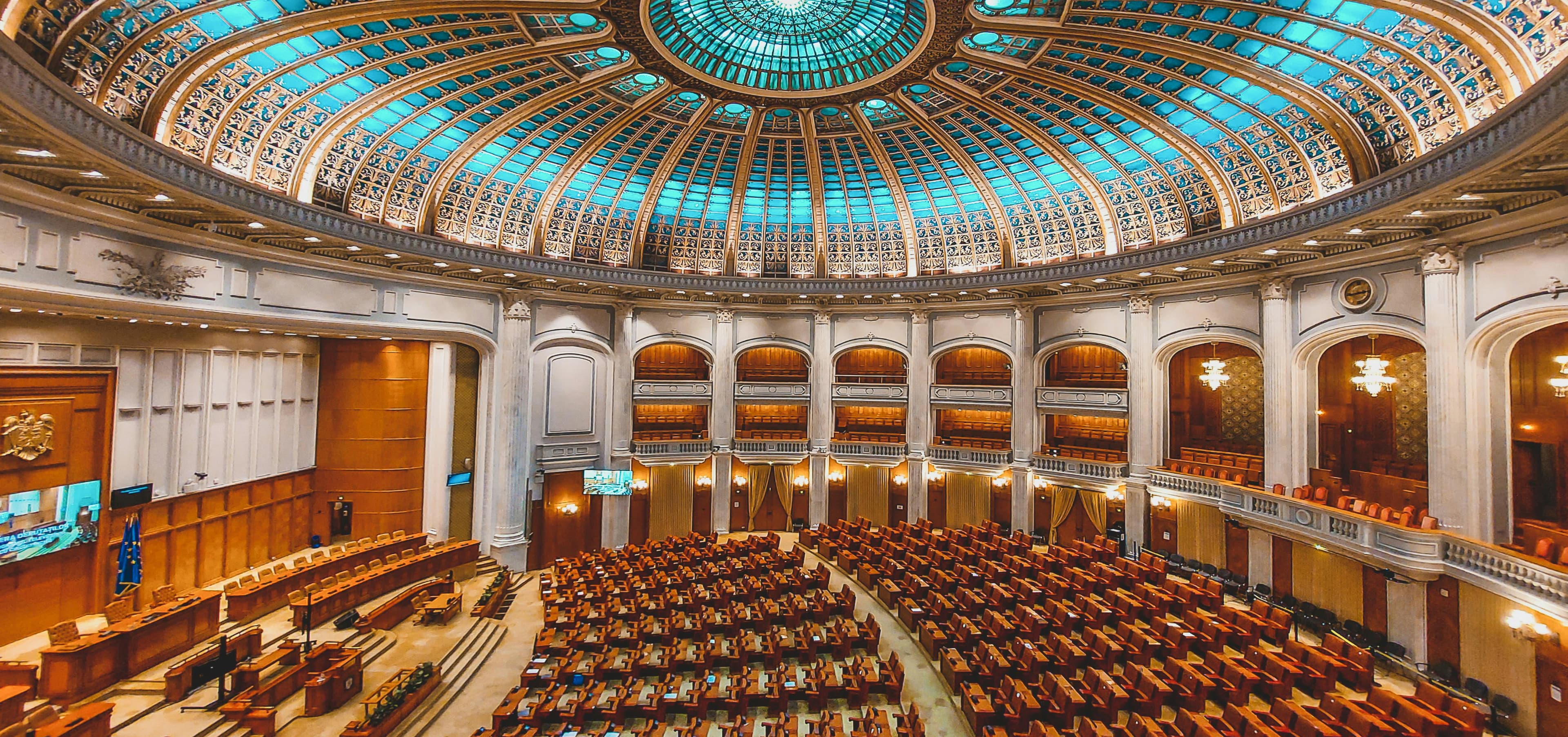 Ornate parliament hall with a blue stained-glass dome ceiling and rows of wooden seats, exuding grandeur.