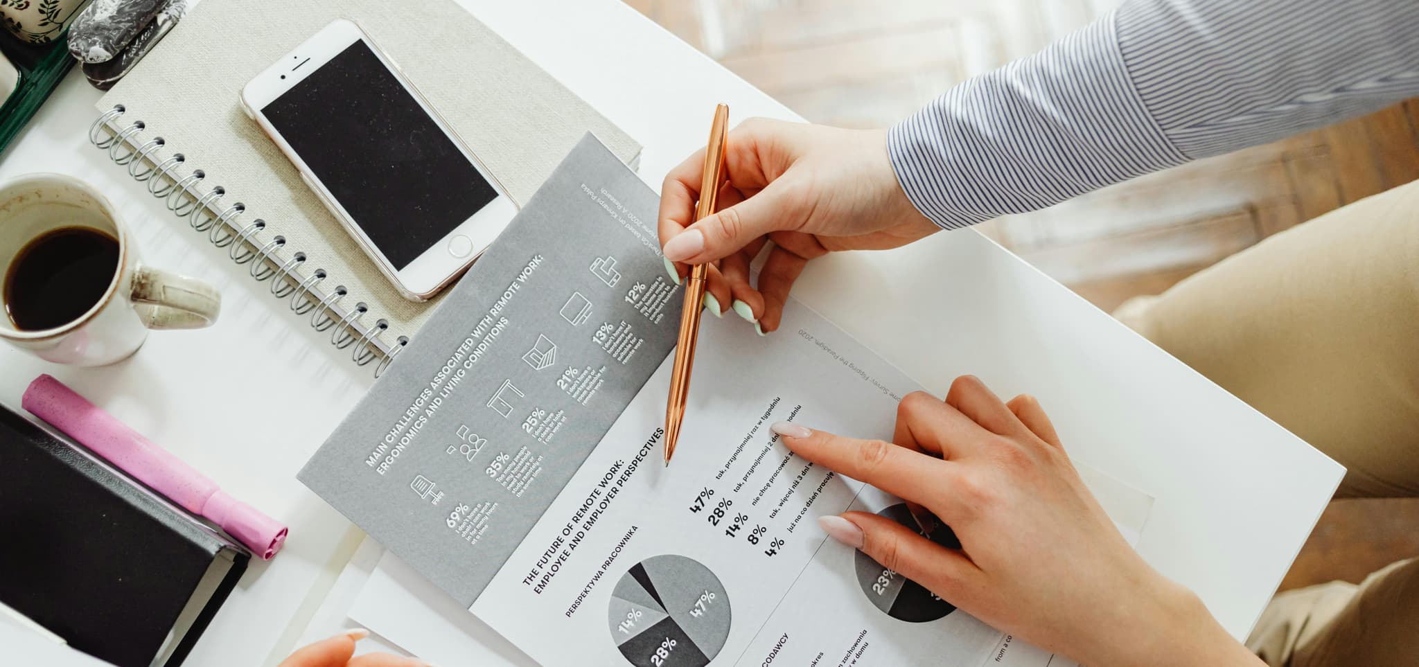 Hands hold a pen, pointing at a document with charts on remote work, with a phone, notebook, coffee, and highlighter.