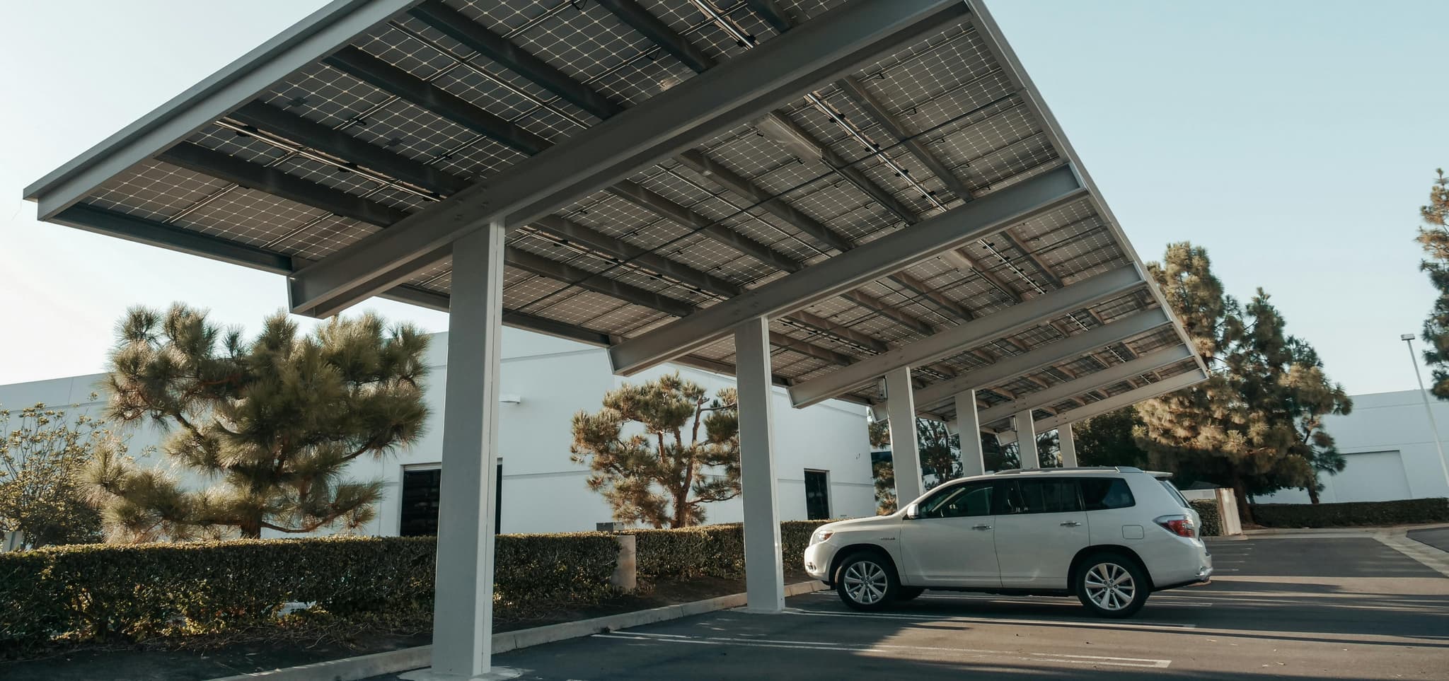 A white SUV parked under solar panel canopies in a sunlit lot, surrounded by green trees, conveying sustainability.