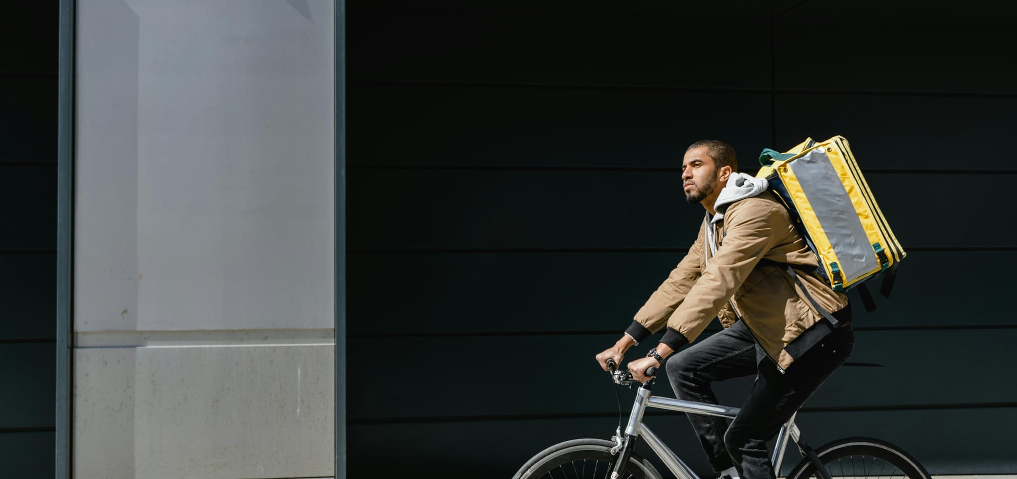 A delivery cyclist in a tan jacket rides past a dark building, carrying a yellow insulated backpack.