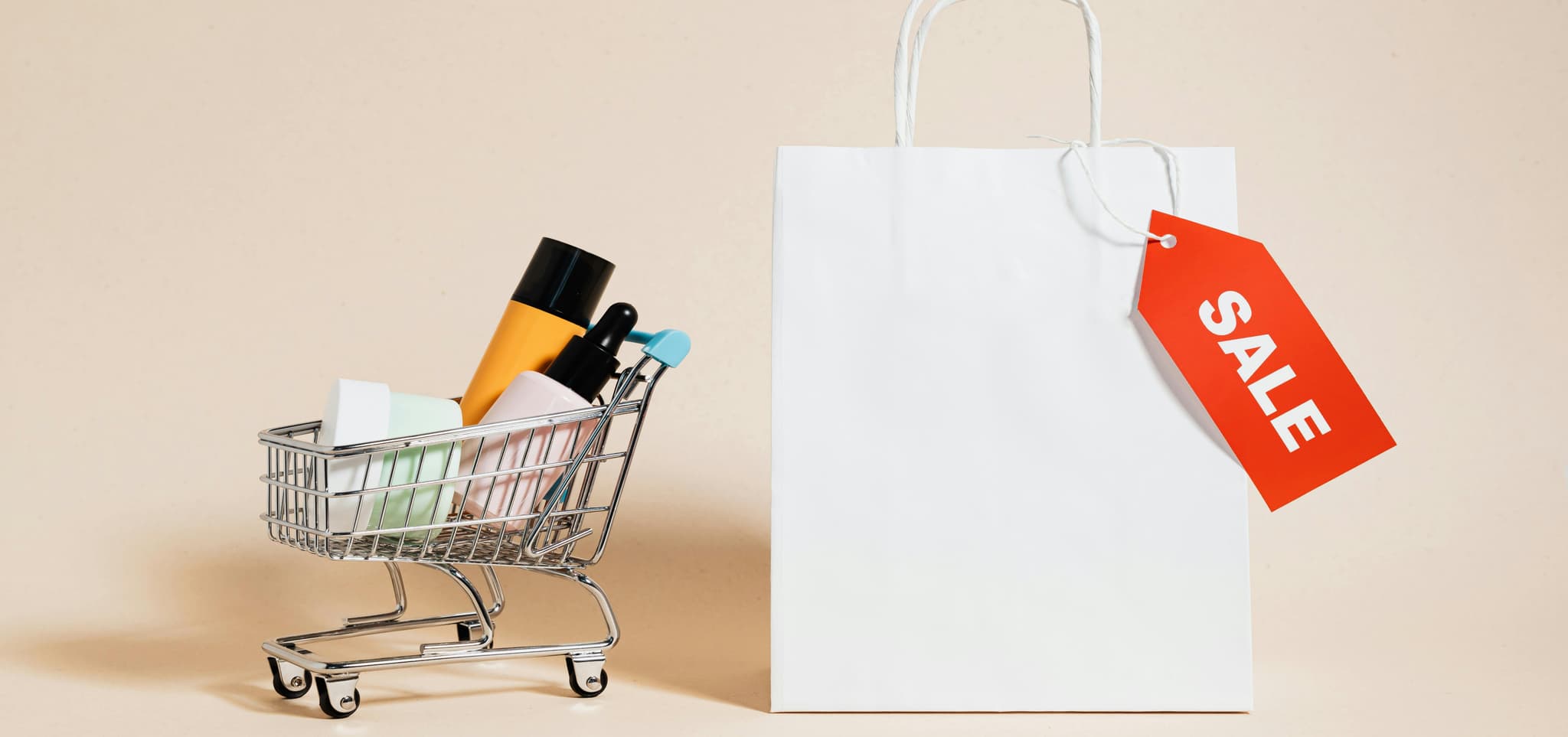 A small shopping cart with colorful bottles next to a white bag with a red 'SALE' tag, symbolizing discount shopping.