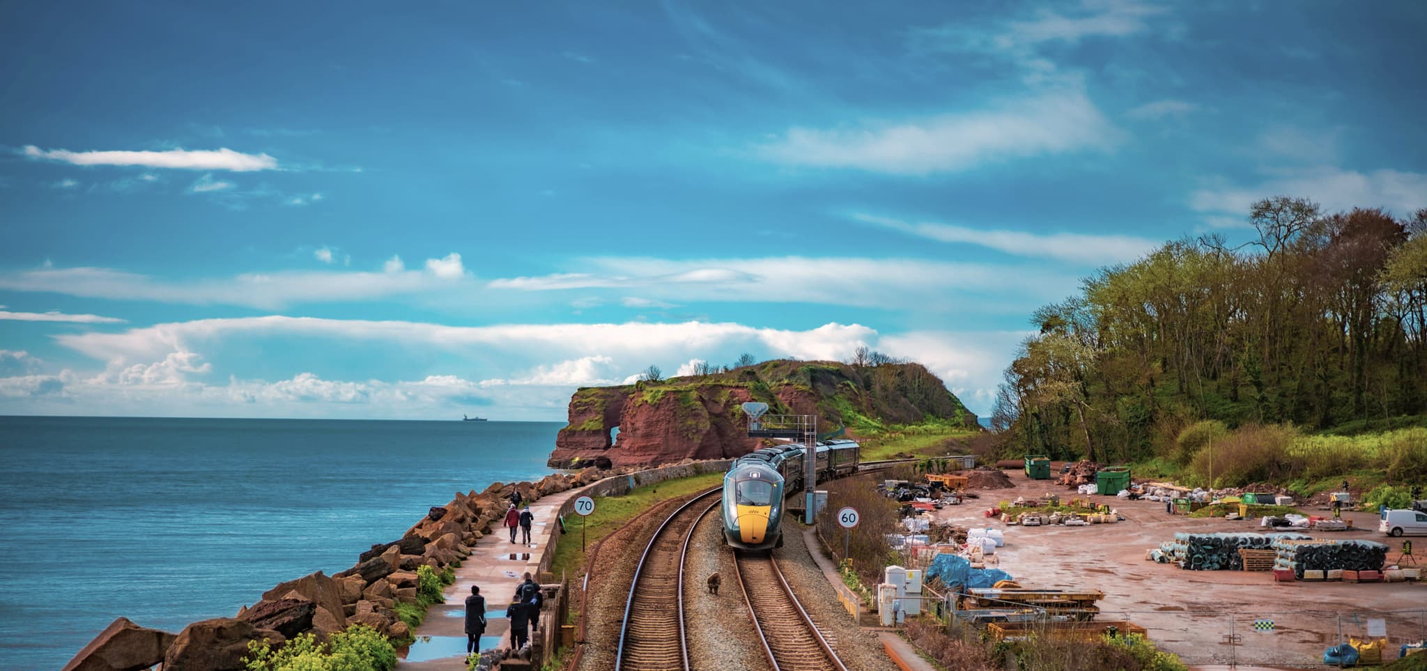 A sleek train winds along coastal tracks with lush greenery, cliffs, and ocean views, as walkers stroll nearby.