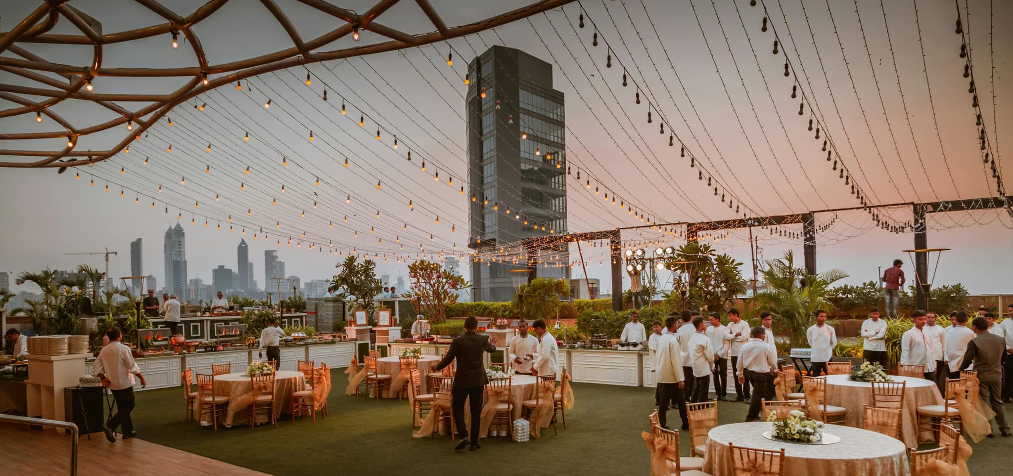 Rooftop event at sunset with elegant tables, string lights, servers in white, and a stunning city skyline.