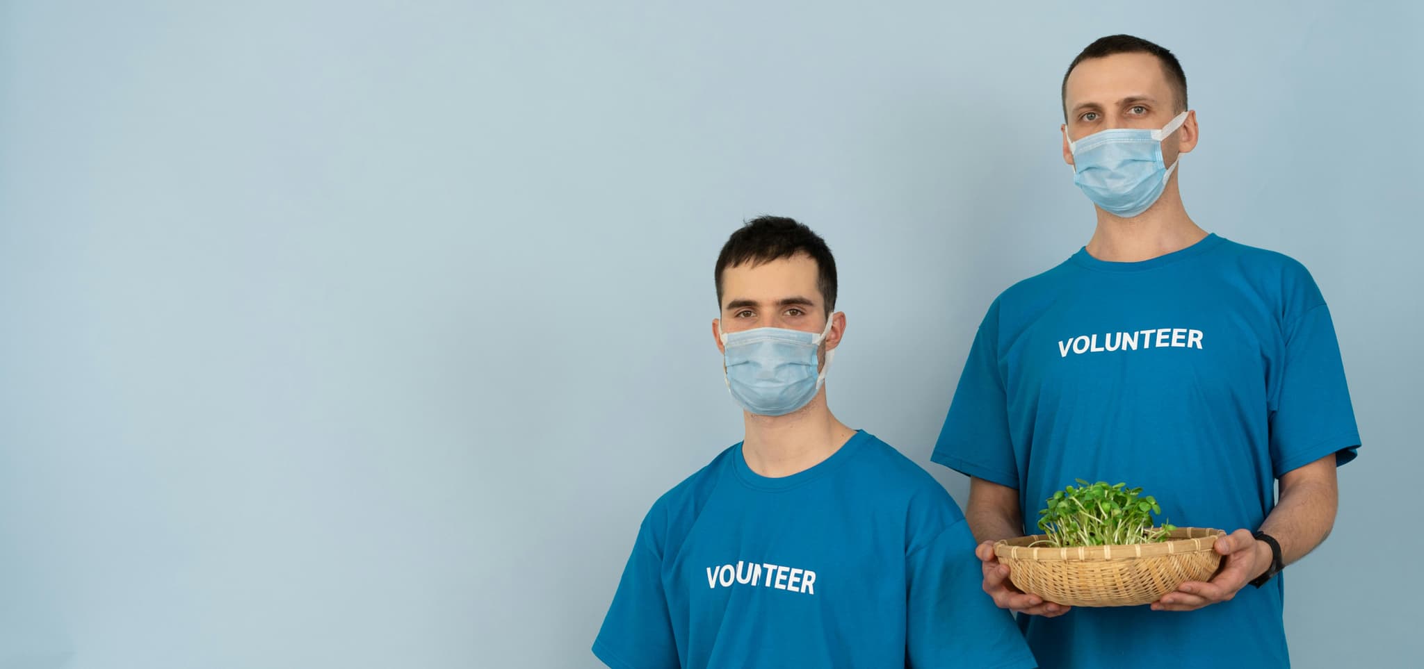 Two masked volunteers in blue shirts, one holding a basket of greens, symbolizing community and care.