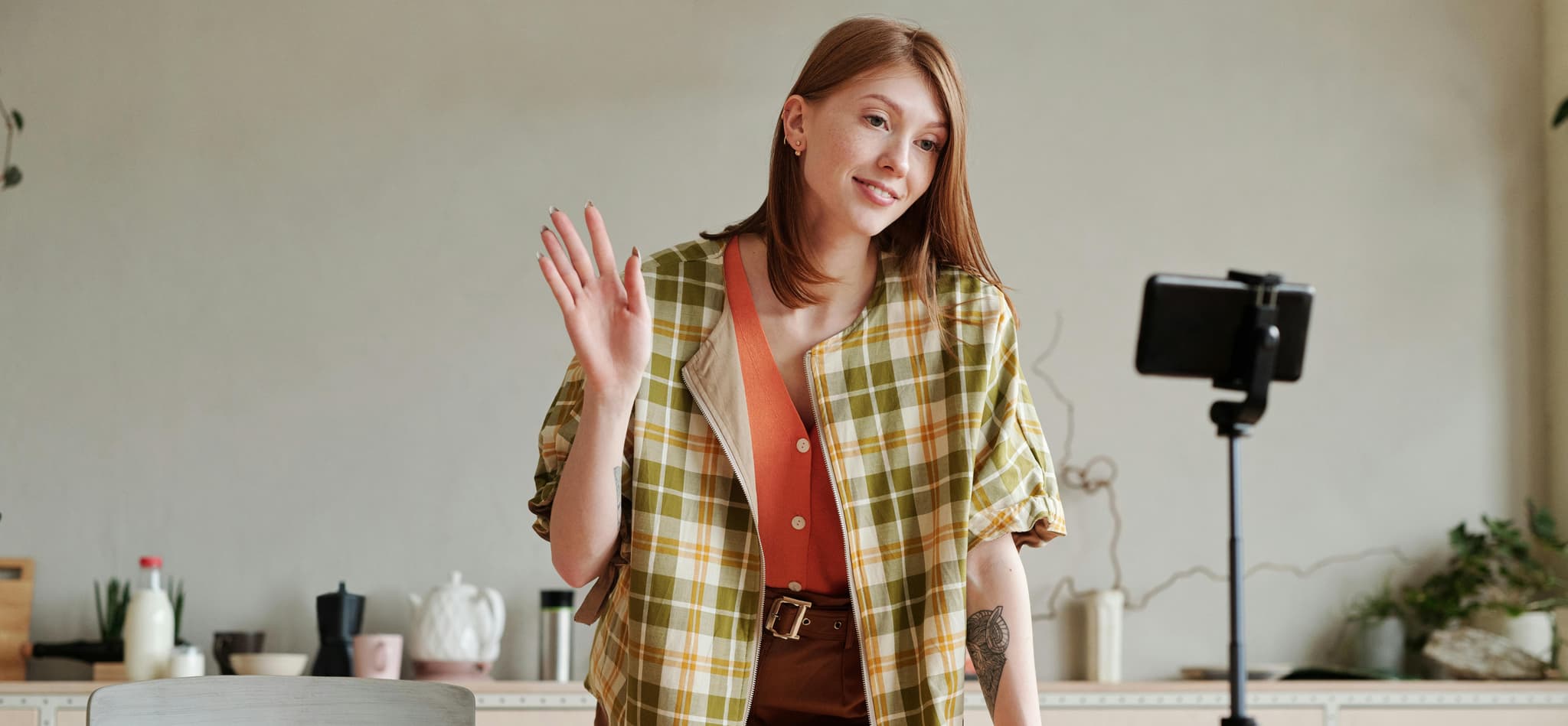 Young woman with red hair smiles and waves at a smartphone on a stand in a cozy, minimal home interior.