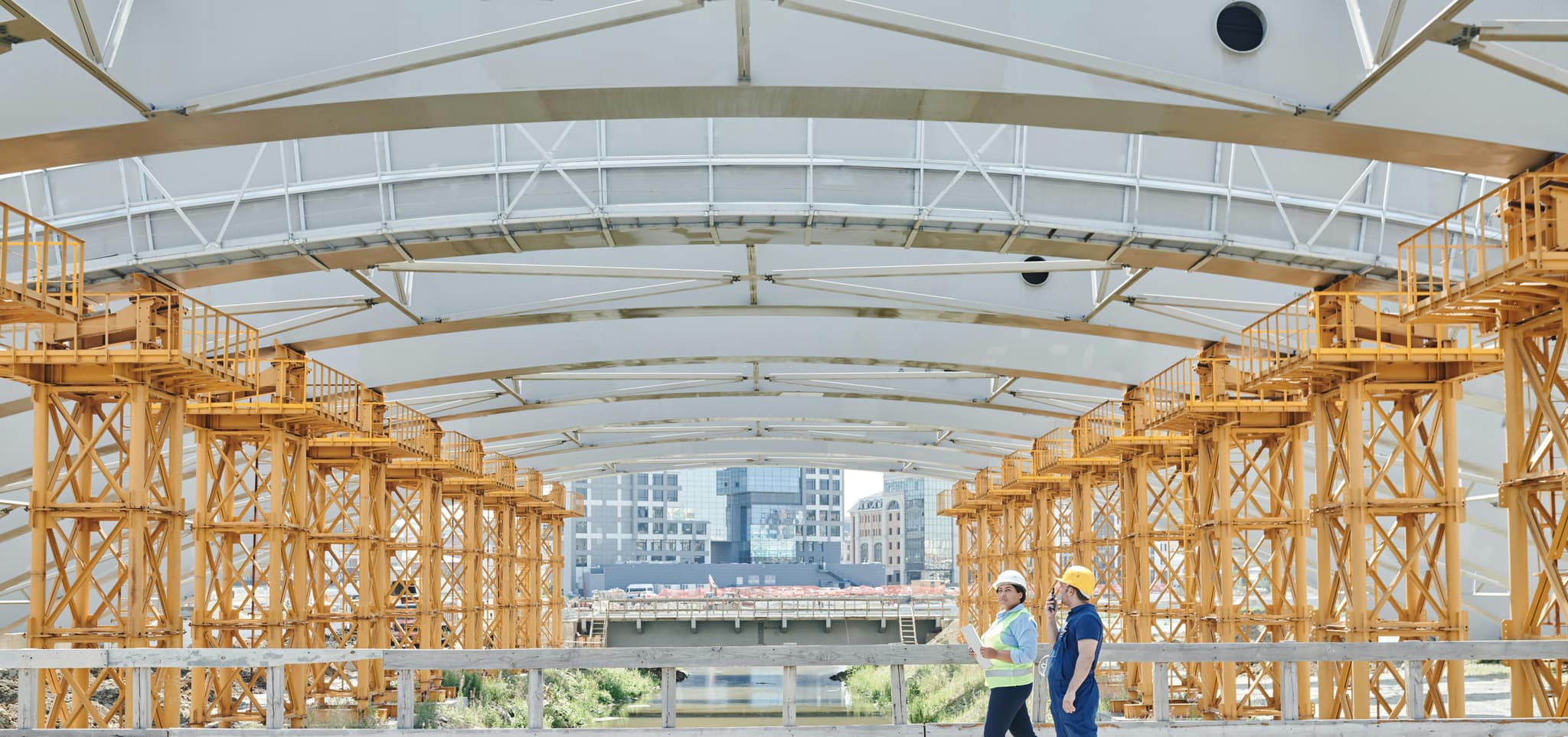 Two construction workers in hard hats and vests walk under a bridge with yellow scaffolding, city skyline behind.