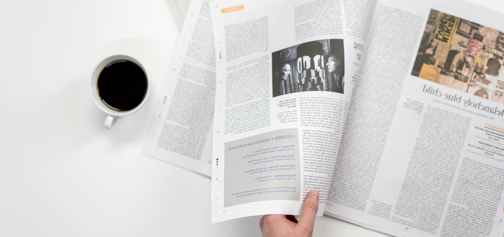 A person holds an open magazine with articles and images, a cup of black coffee on a white table, evoking calm.