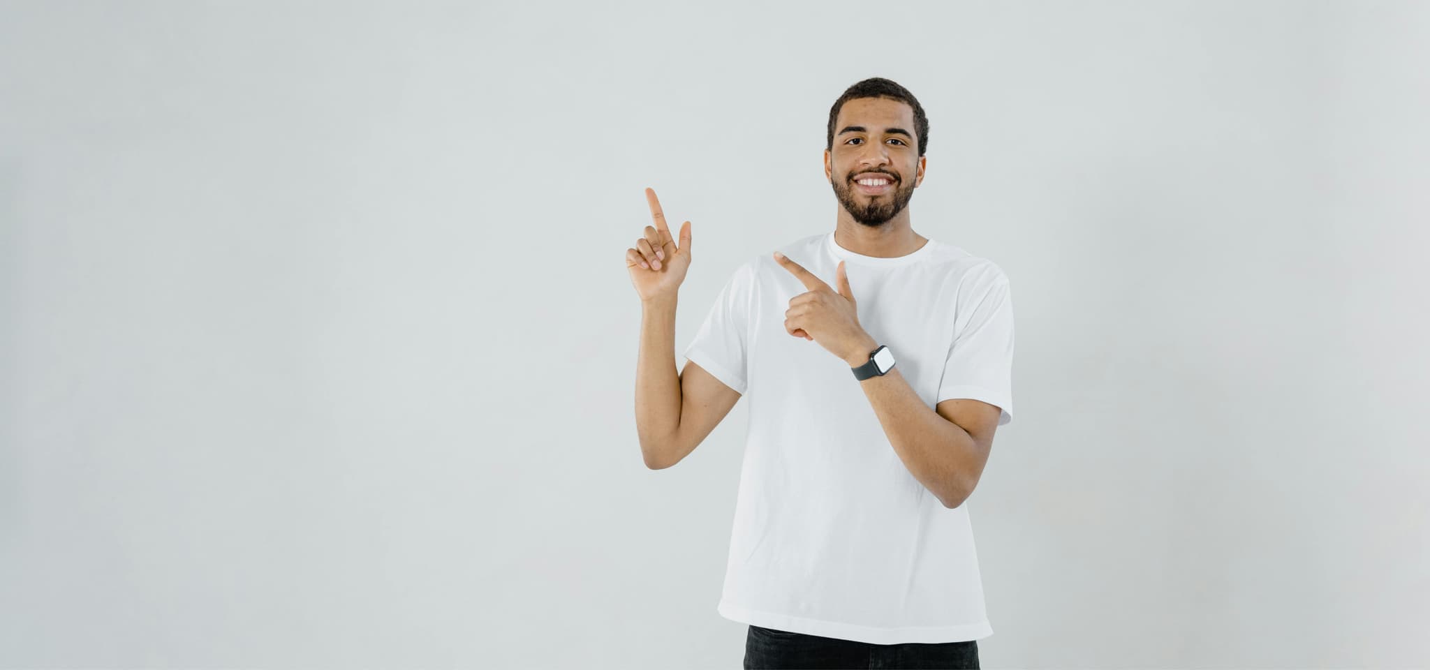 Smiling man in a white T-shirt points up with both hands, wearing a smartwatch against a light gray background.