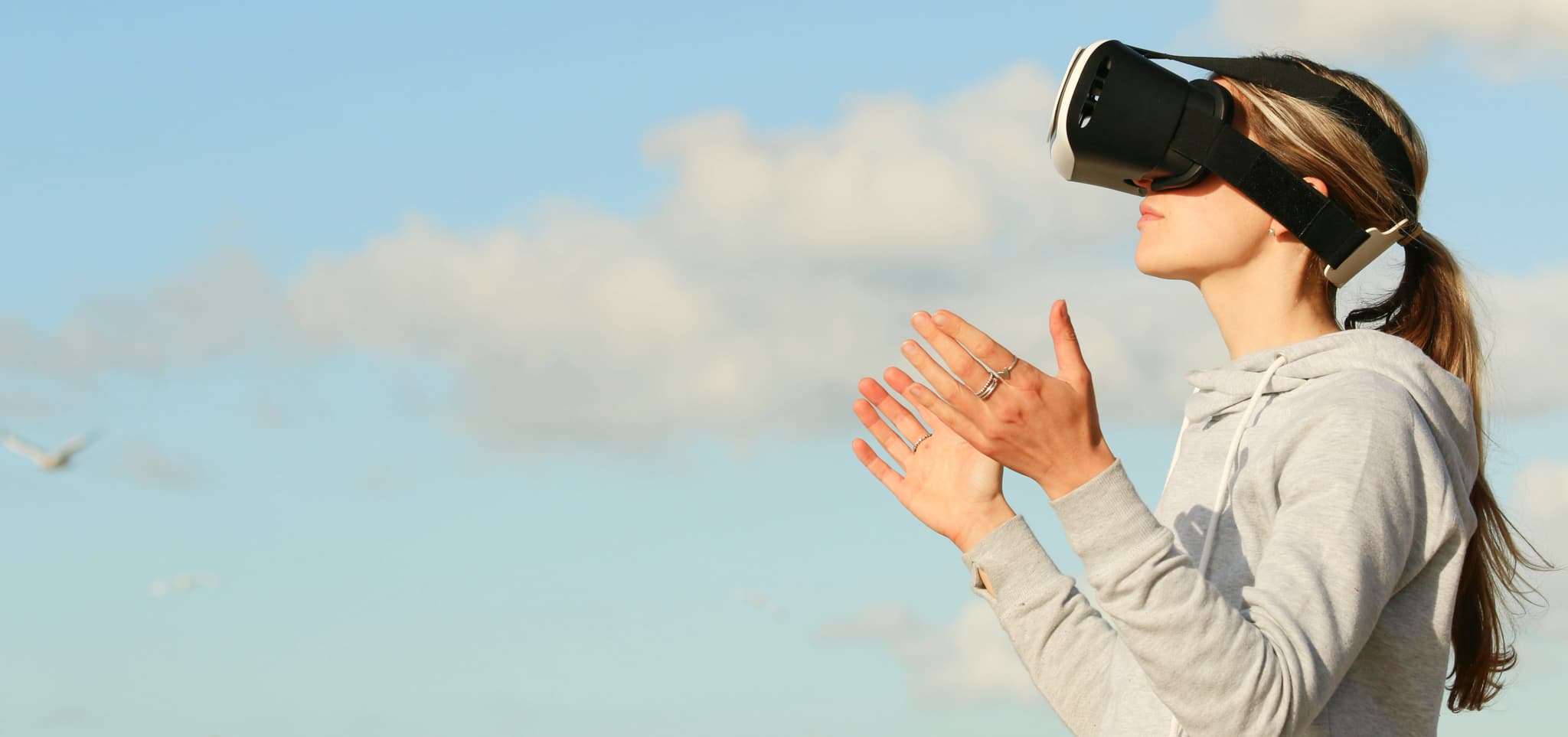 A person in a gray hoodie wearing a VR headset, clapping hands under a blue sky with clouds, evoking wonder.